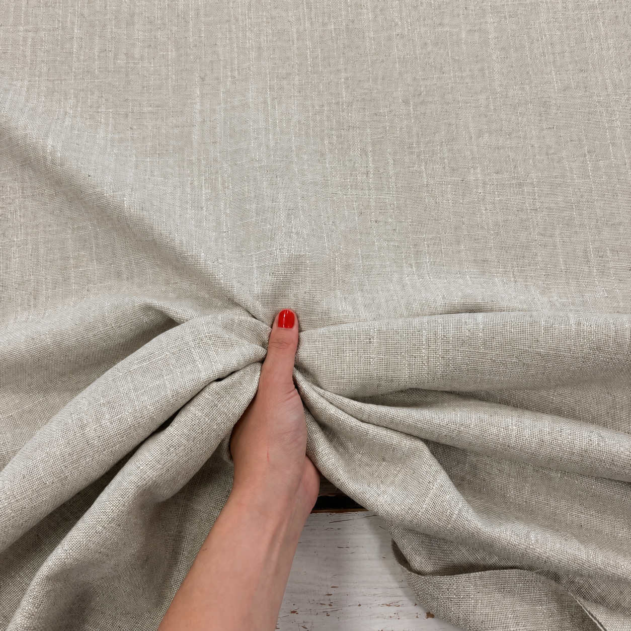 A close-up of light beige textured fabric being gathered and pulled by a hand with red nail polish, showing the material's weave and drape.