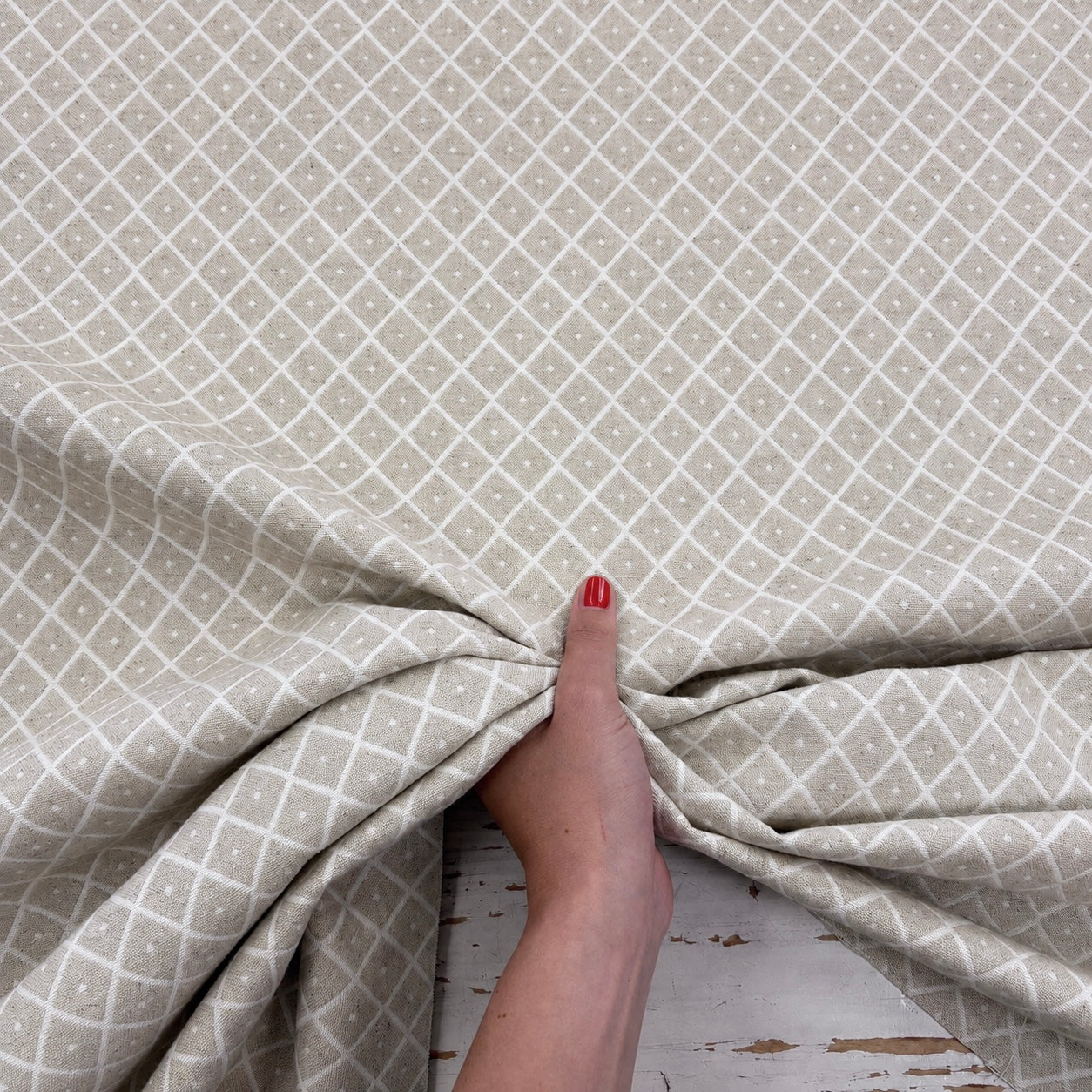 A close-up of a person's hand with red nail polish pulling a piece of light beige fabric with a white geometric diamond pattern.