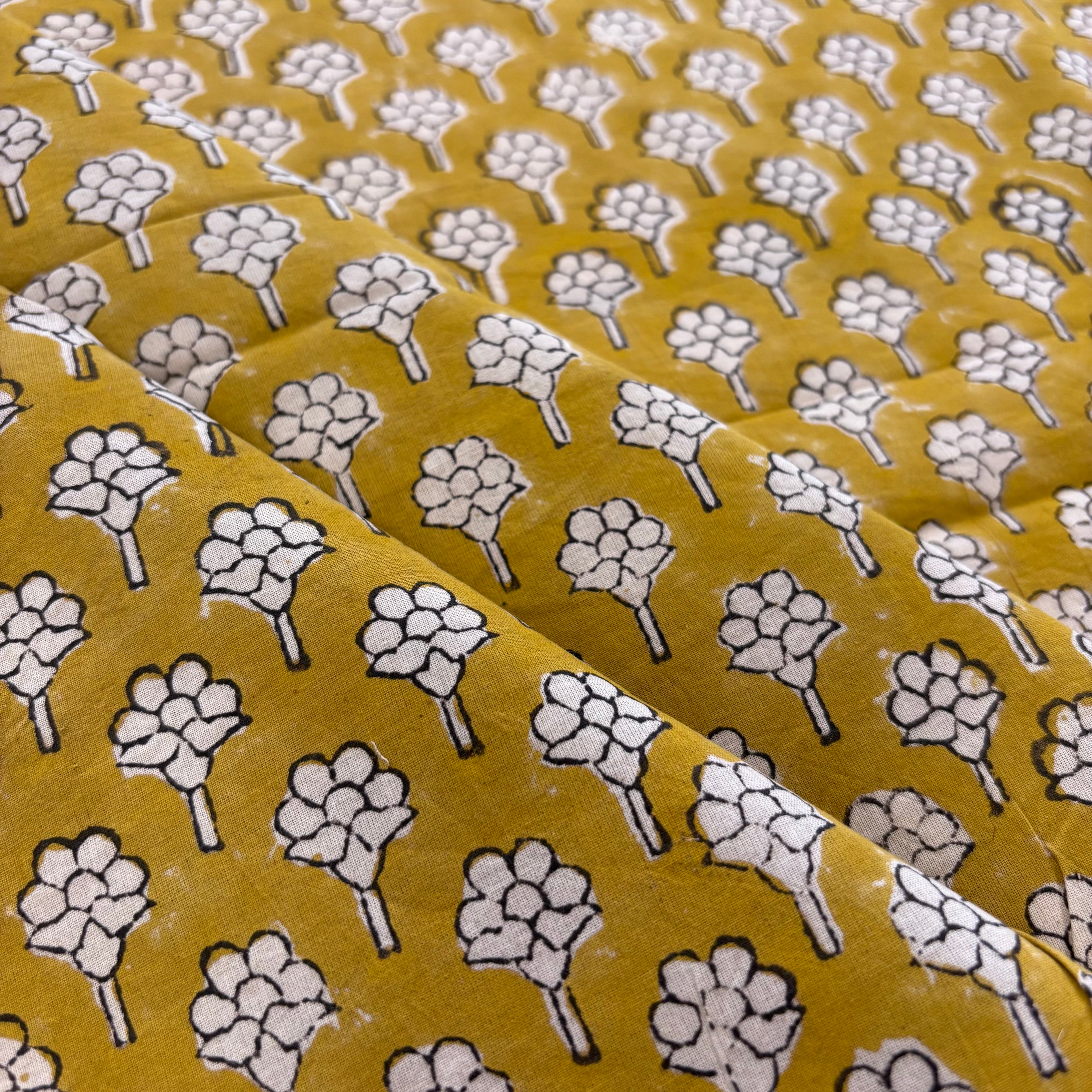 A close-up of a mustard yellow fabric with a repeating white and black block-printed floral pattern, showing soft folds and texture.