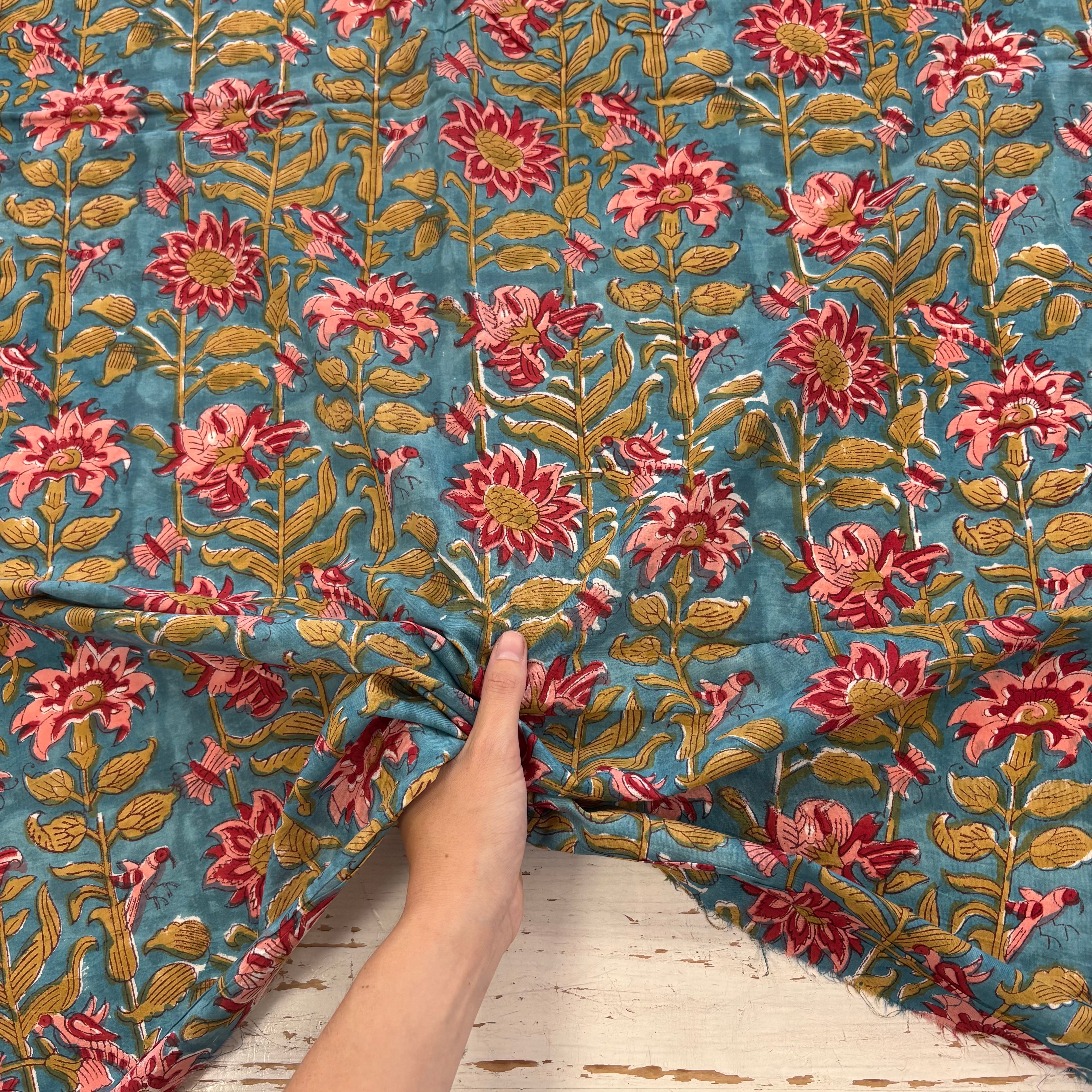 A close-up of a hand holding a piece of blue cotton fabric with a detailed floral block print in pink, red, and green, set against a rustic white wooden background.
