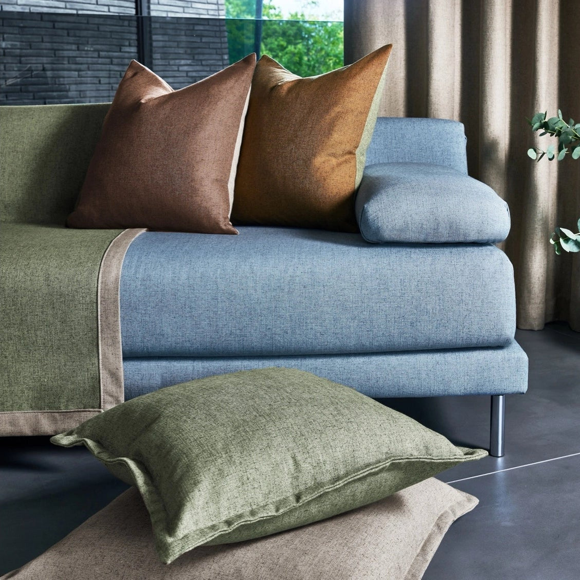 Blue sofa with brown and green cushions in a room with curtains and a plant.