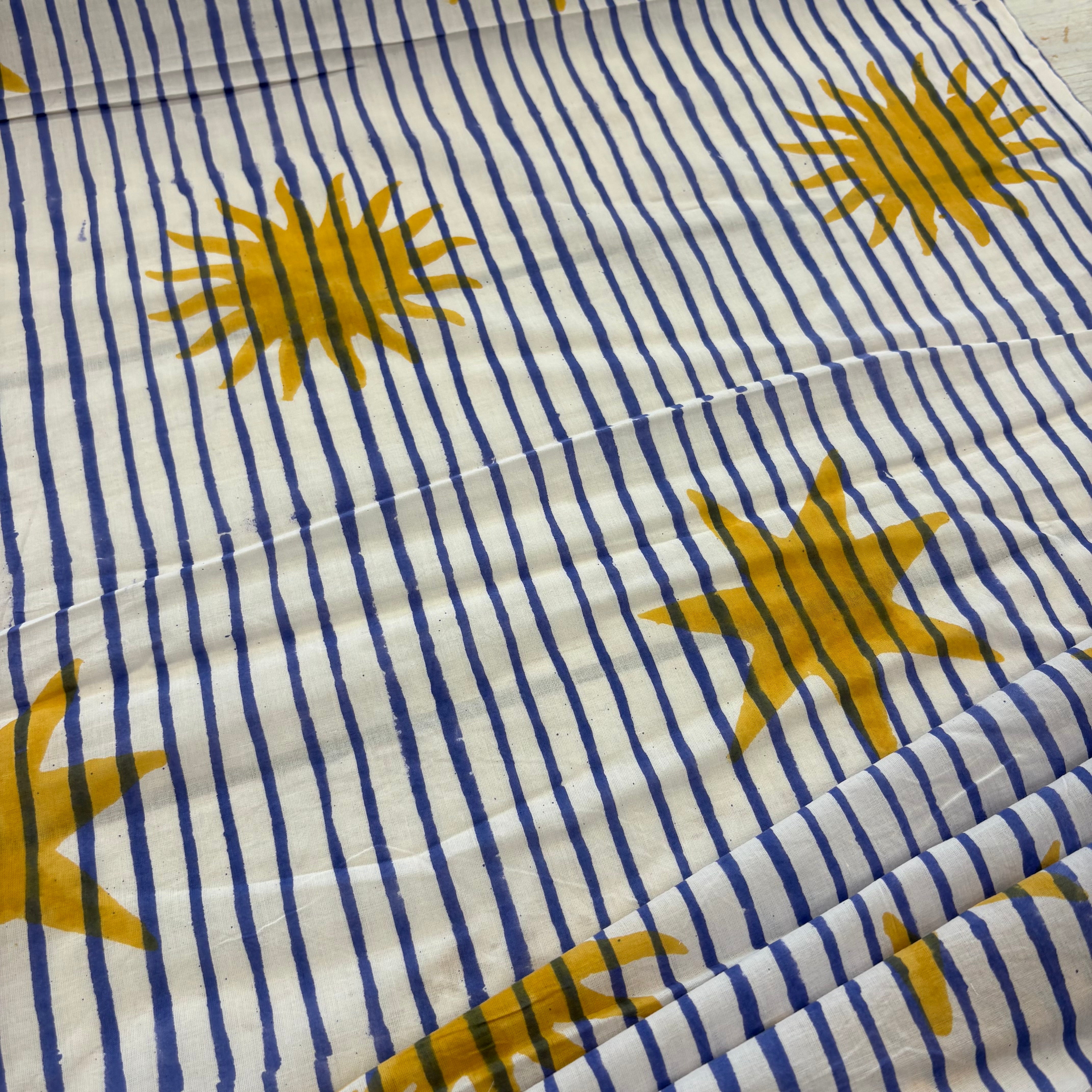A close-up of a fabric surface with vertical navy blue stripes on an off-white background and large mustard-yellow sun motifs scattered across it.
