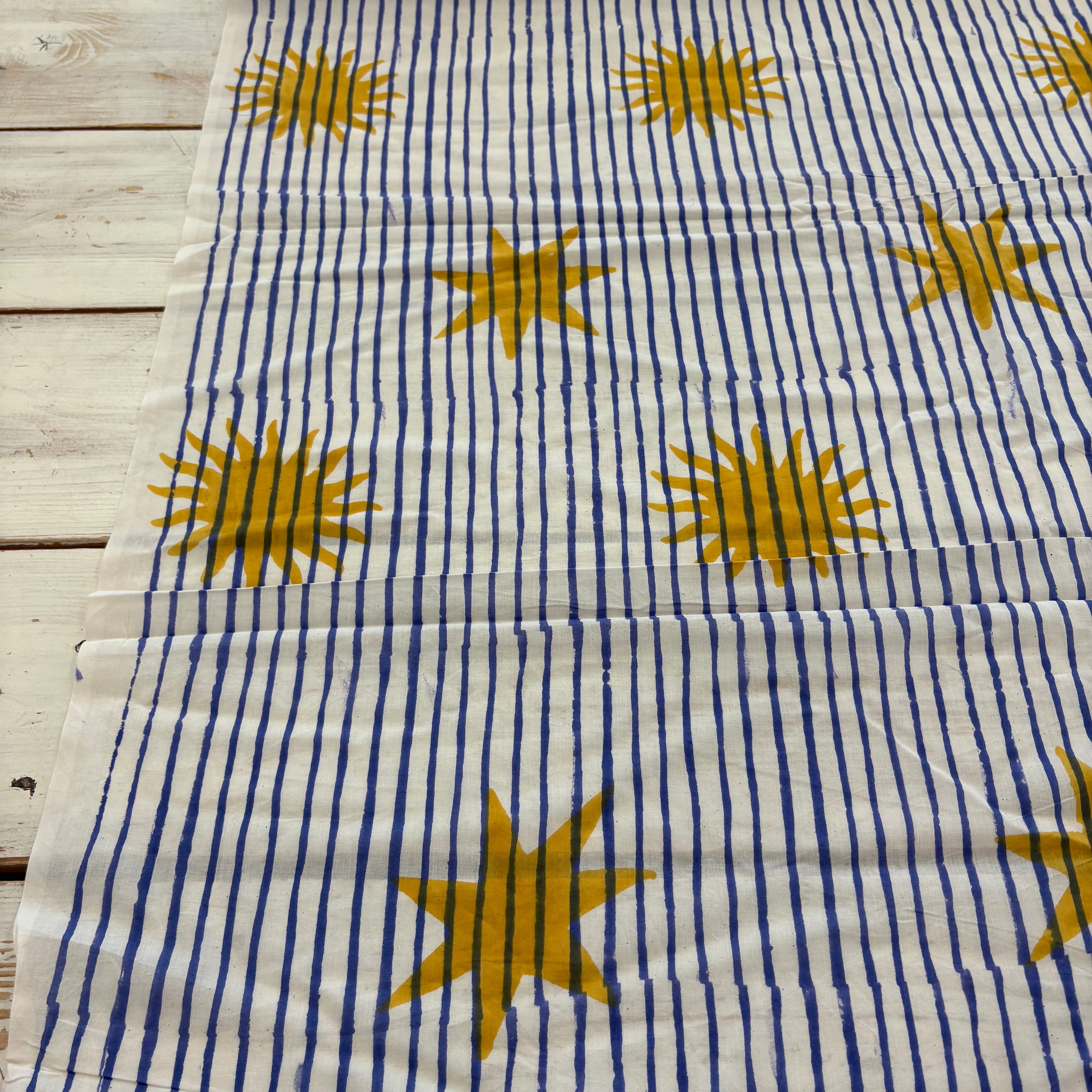 A fabric laid flat featuring a pattern of vertical blue stripes and large mustard yellow starburst motifs, displayed on a light-colored wooden surface.