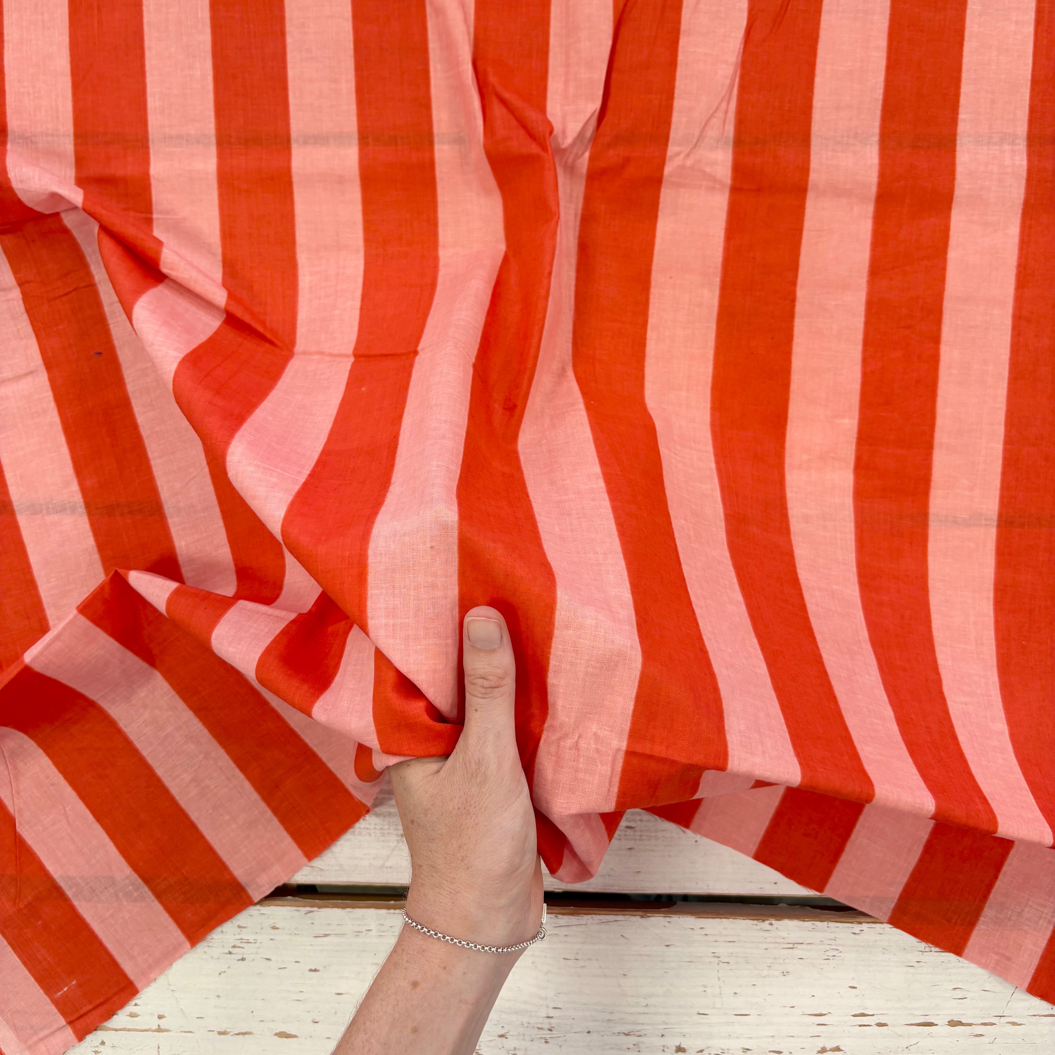 A close-up of a hand holding a piece of fabric with vertical stripes in bright orange-red and light pink, draped over a distressed white wooden surface.