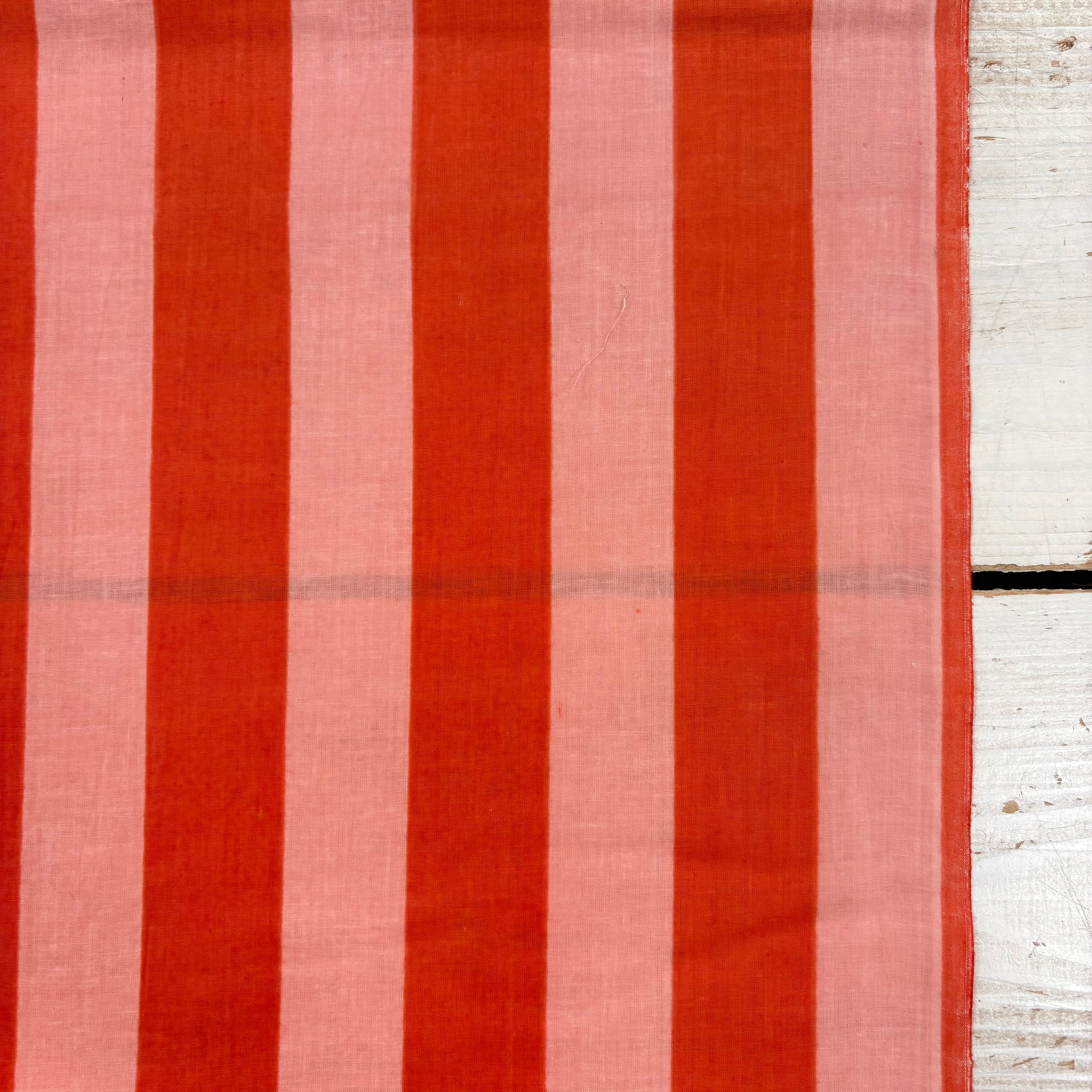 A close-up of a striped textile with alternating vertical bands of rust red and soft pink, laid flat against a white wooden surface.