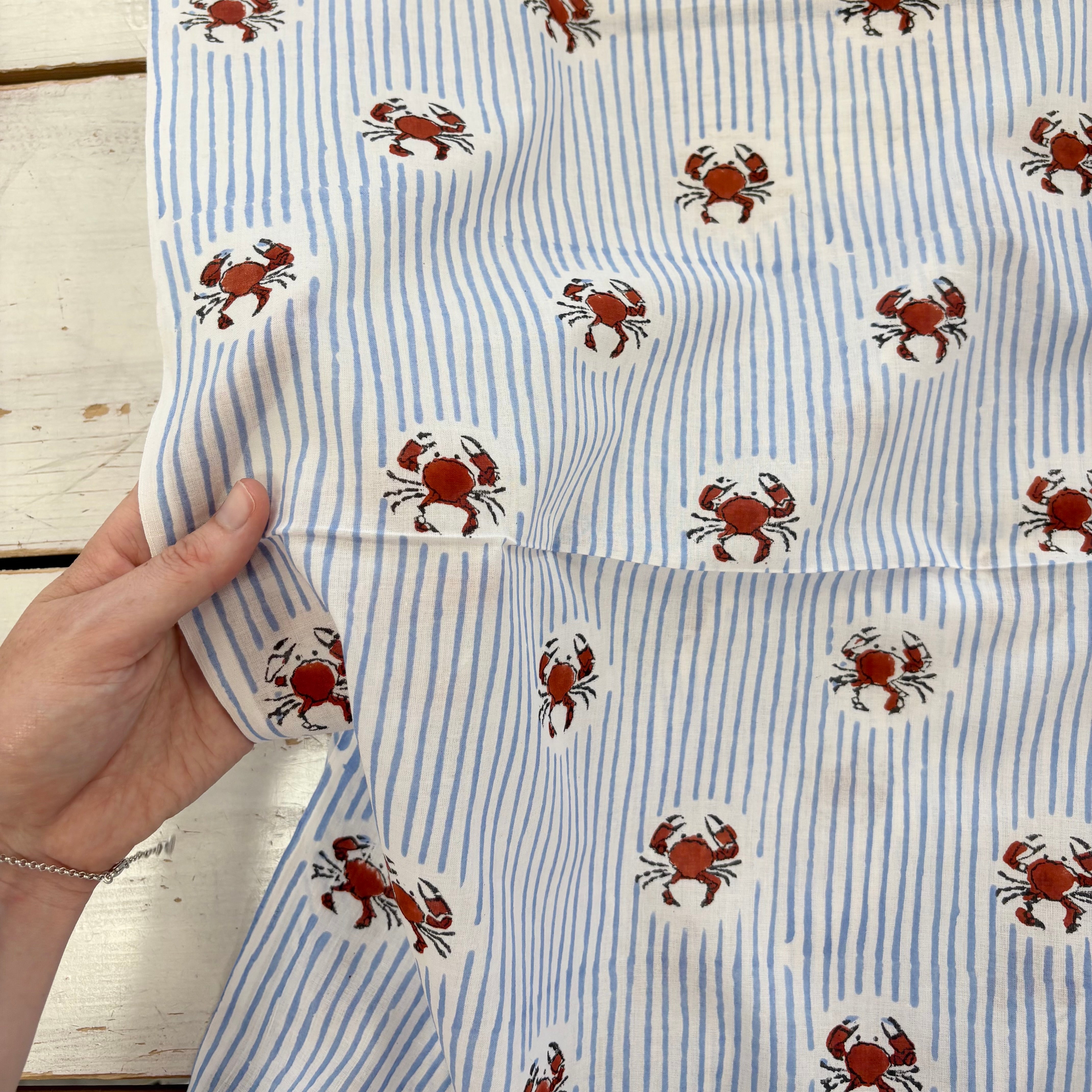 A close-up of a hand holding a piece of fabric with a pattern of blue and white vertical stripes and red crabs on a white background, laid over a white wooden surface.
