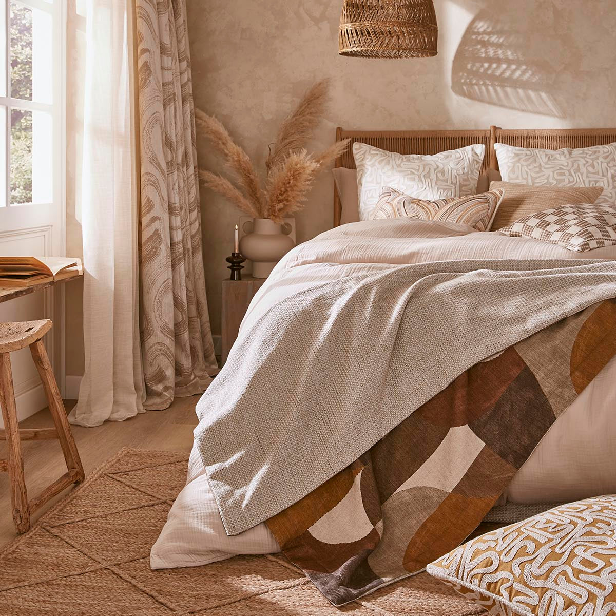 A cozy bedroom scene with a bed dressed in neutral-toned bedding, including a cream duvet and patterned pillows, styled with a woven pendant light, textured wallpaper, and a window with sheer curtains.