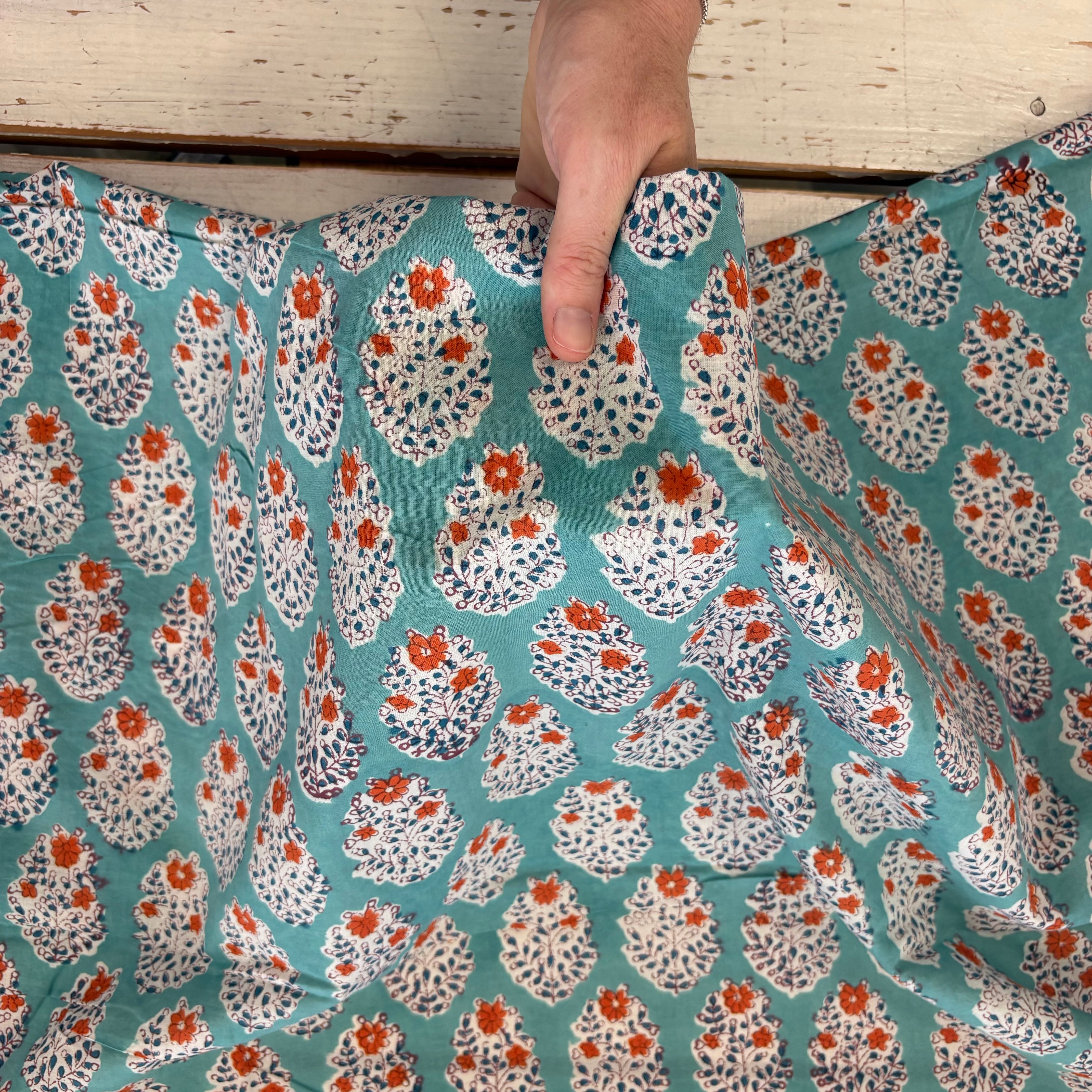 A close-up of a teal fabric with a repeating white, orange, and dark blue floral block print pattern, held up by a hand against a rustic light wood background.