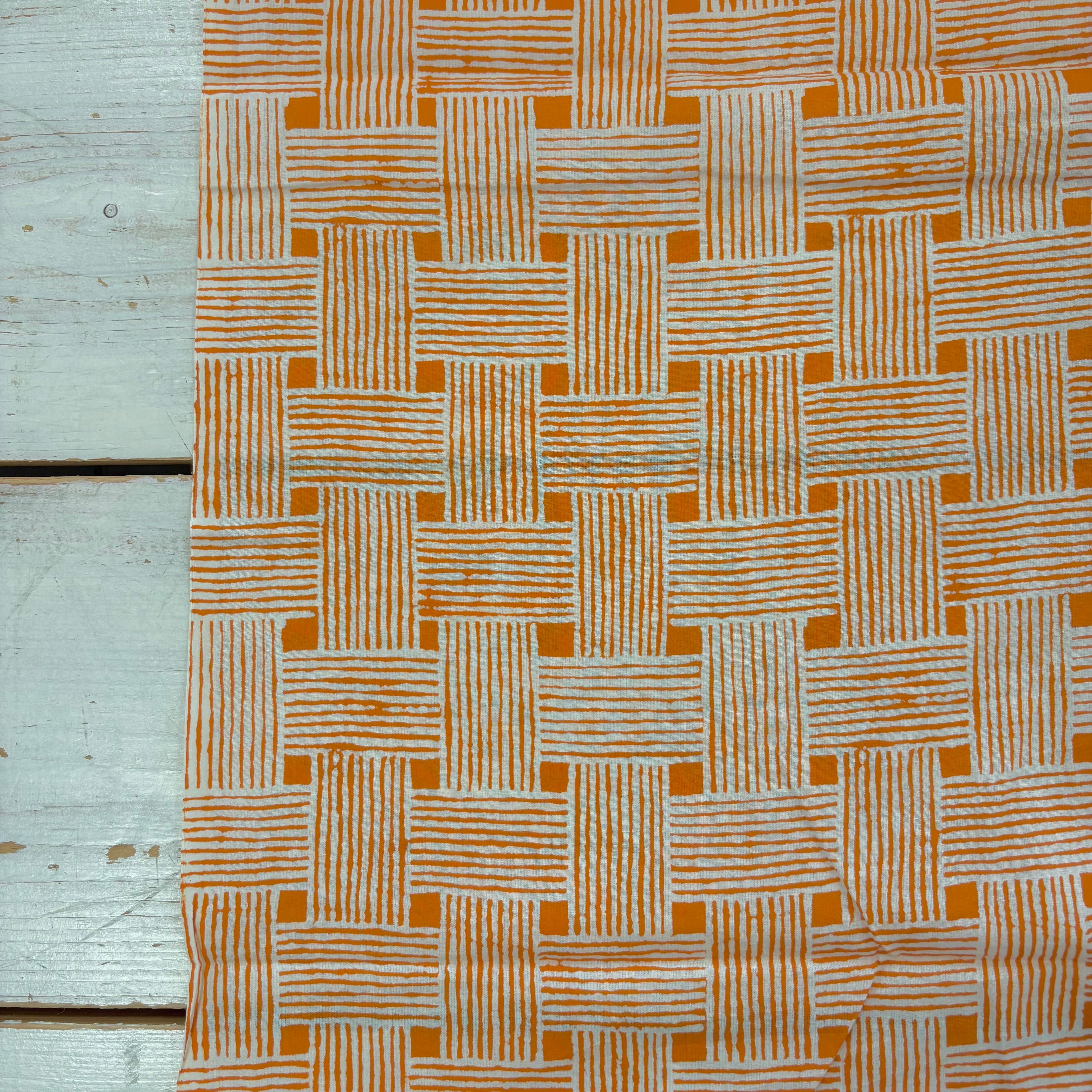 A close-up of a woven basketweave fabric pattern in orange and off-white, laid flat on a light gray wooden surface.