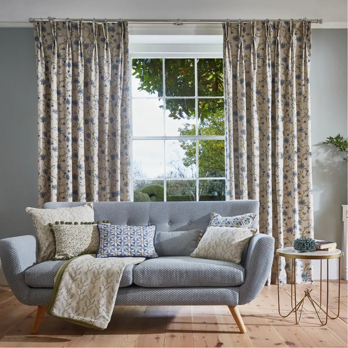 A styled living room scene featuring a light gray tufted sofa with patterned throw pillows and a cream throw blanket, positioned in front of a large window with matching floral curtains. The room has light gray walls and a light wood floor.