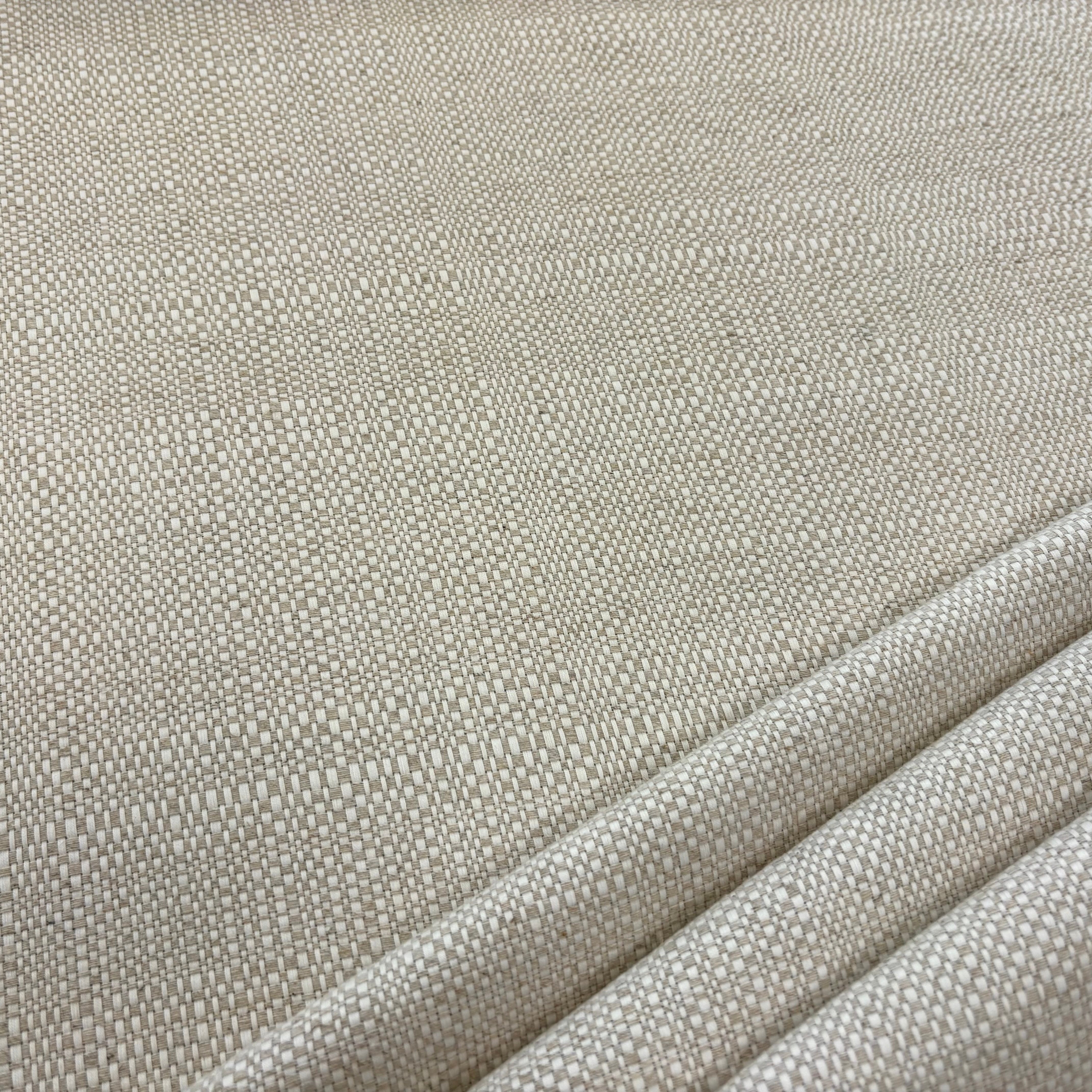 A close-up of a woven fabric with a textured, basketweave-like pattern in a light beige or oatmeal color, with soft diagonal folds visible on the bottom right.