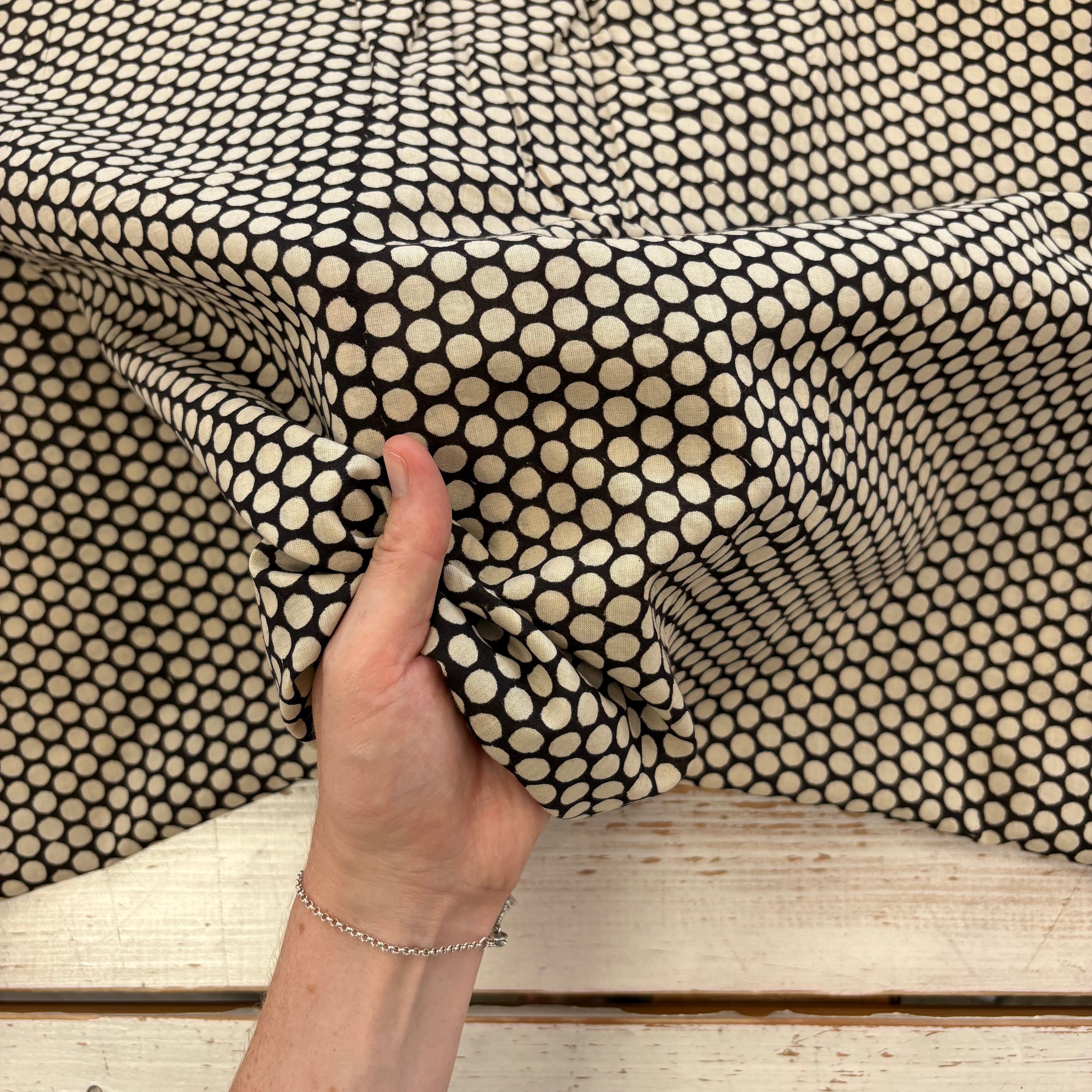 A close-up of a hand holding up a piece of fabric with a black and cream geometric dot pattern, draped over a rustic white wooden surface.