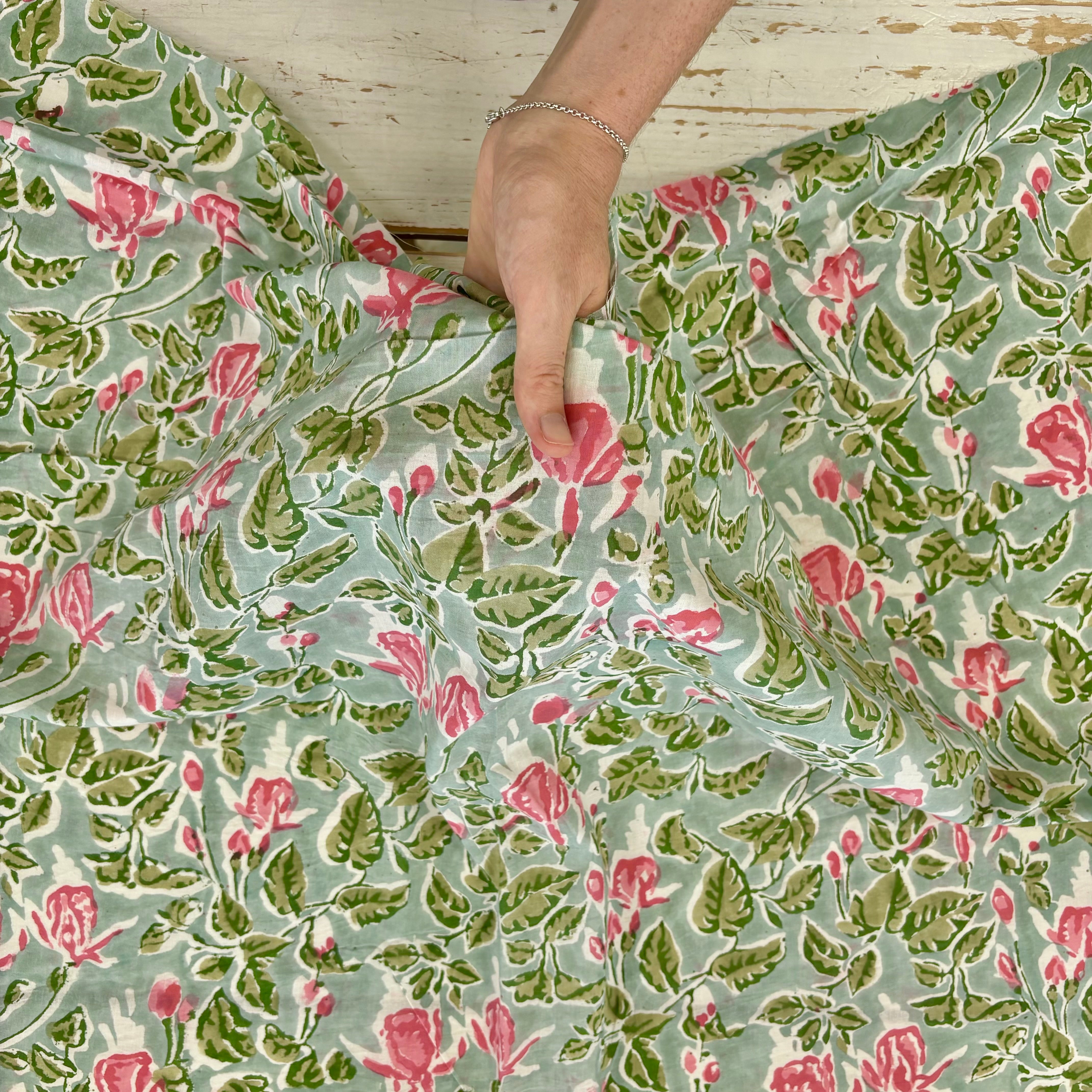 A close-up of a floral fabric with a sage green background, pink roses, and green leaves, held by a hand over a rustic white wooden surface.
