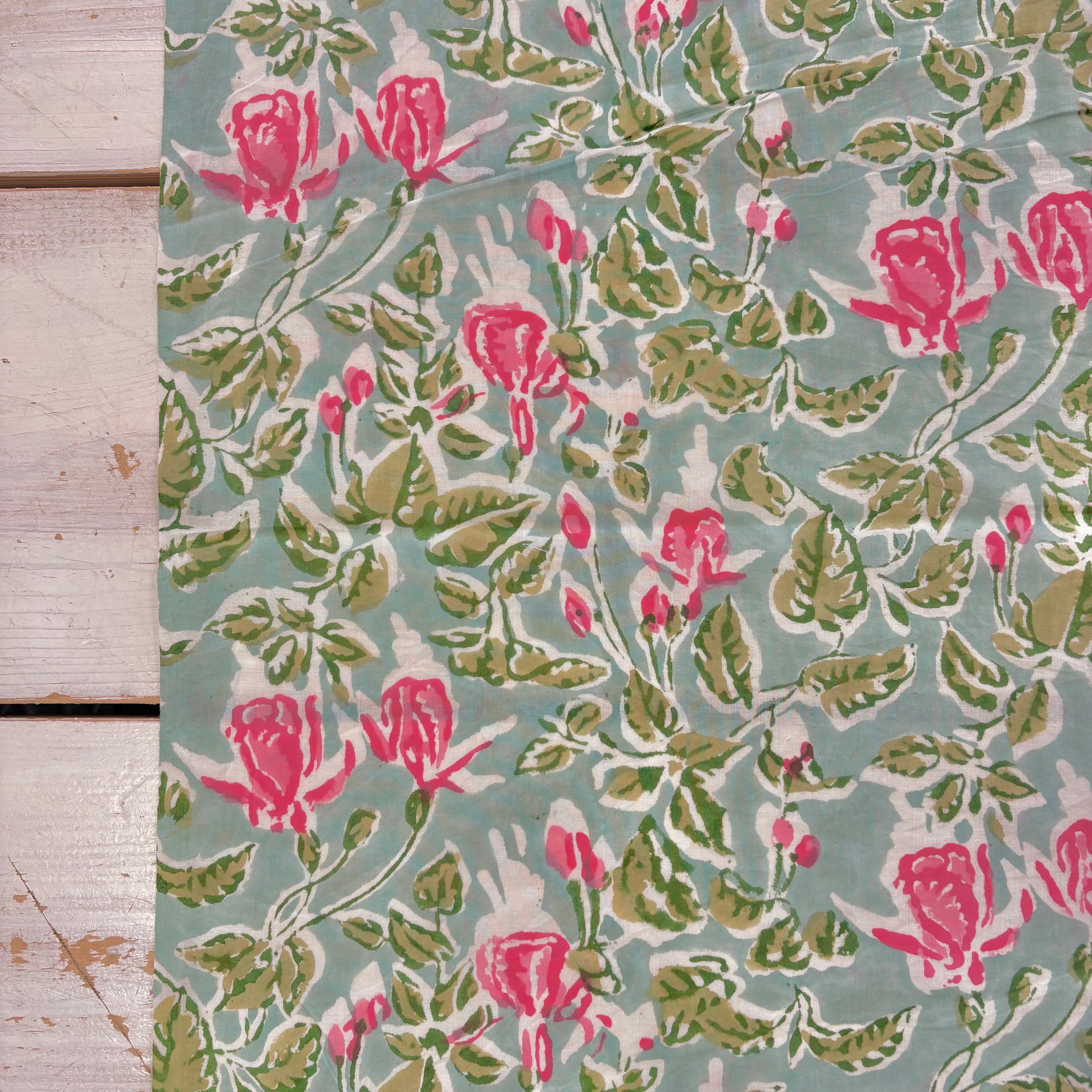 A close-up of a floral fabric or wallpaper pattern with pink roses and green leaves on a muted sage background, laid flat next to a light wood surface.