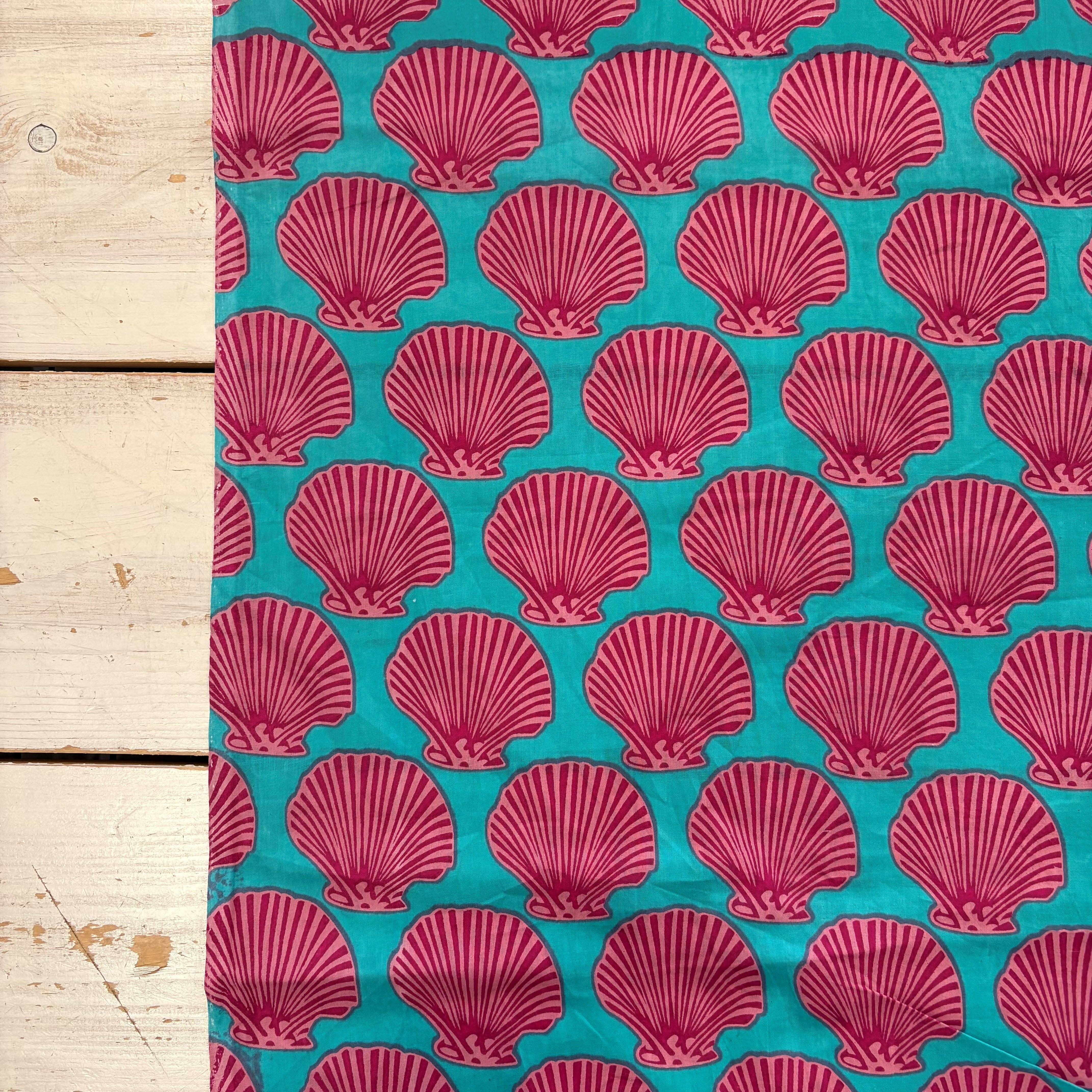 A close-up of a fabric pattern featuring repeating magenta-pink scallop shell motifs on a bright turquoise background, laid next to a light wood plank surface.