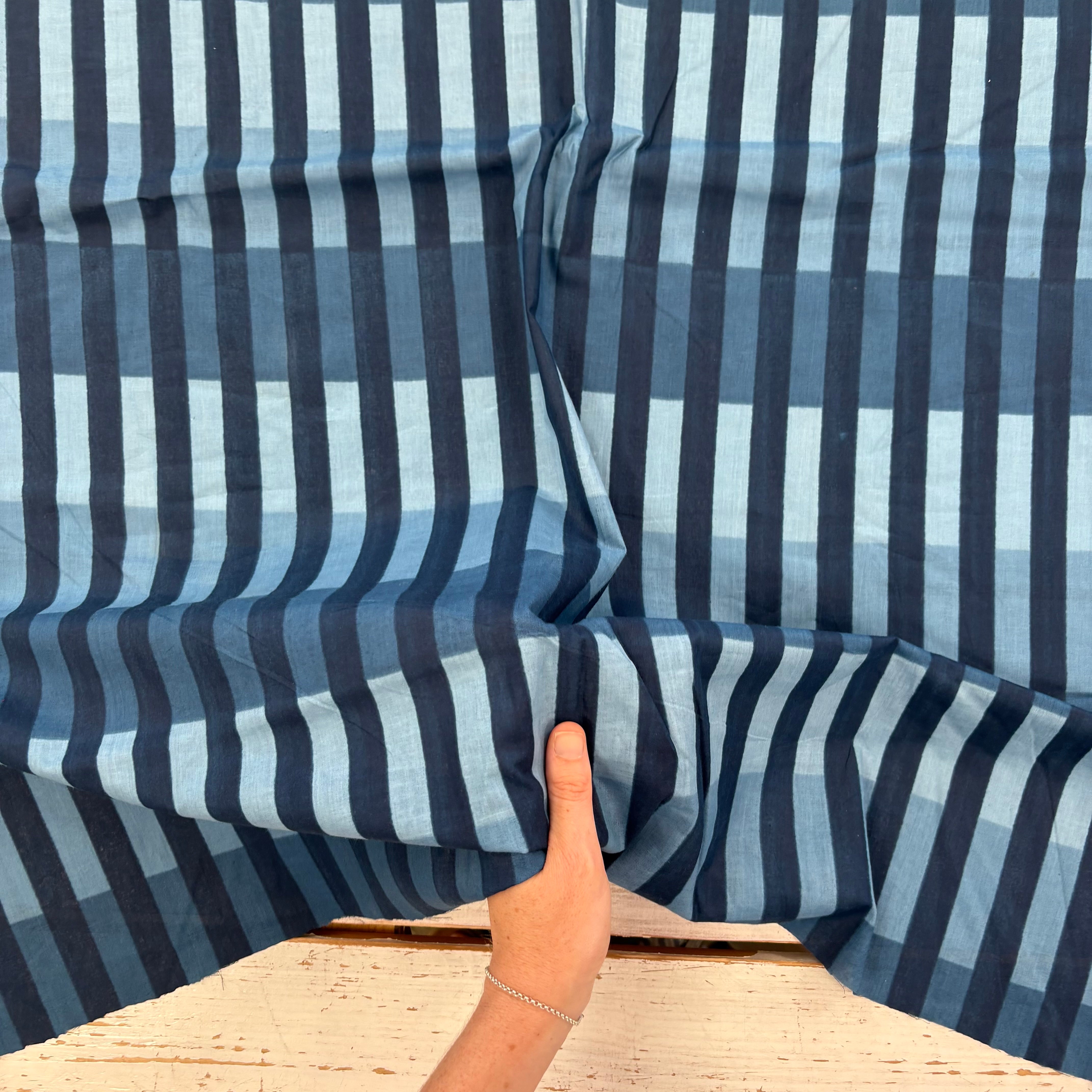 A close-up of a striped fabric with vertical bands of navy, medium blue, and light blue, held up by a hand to show its texture and pattern.