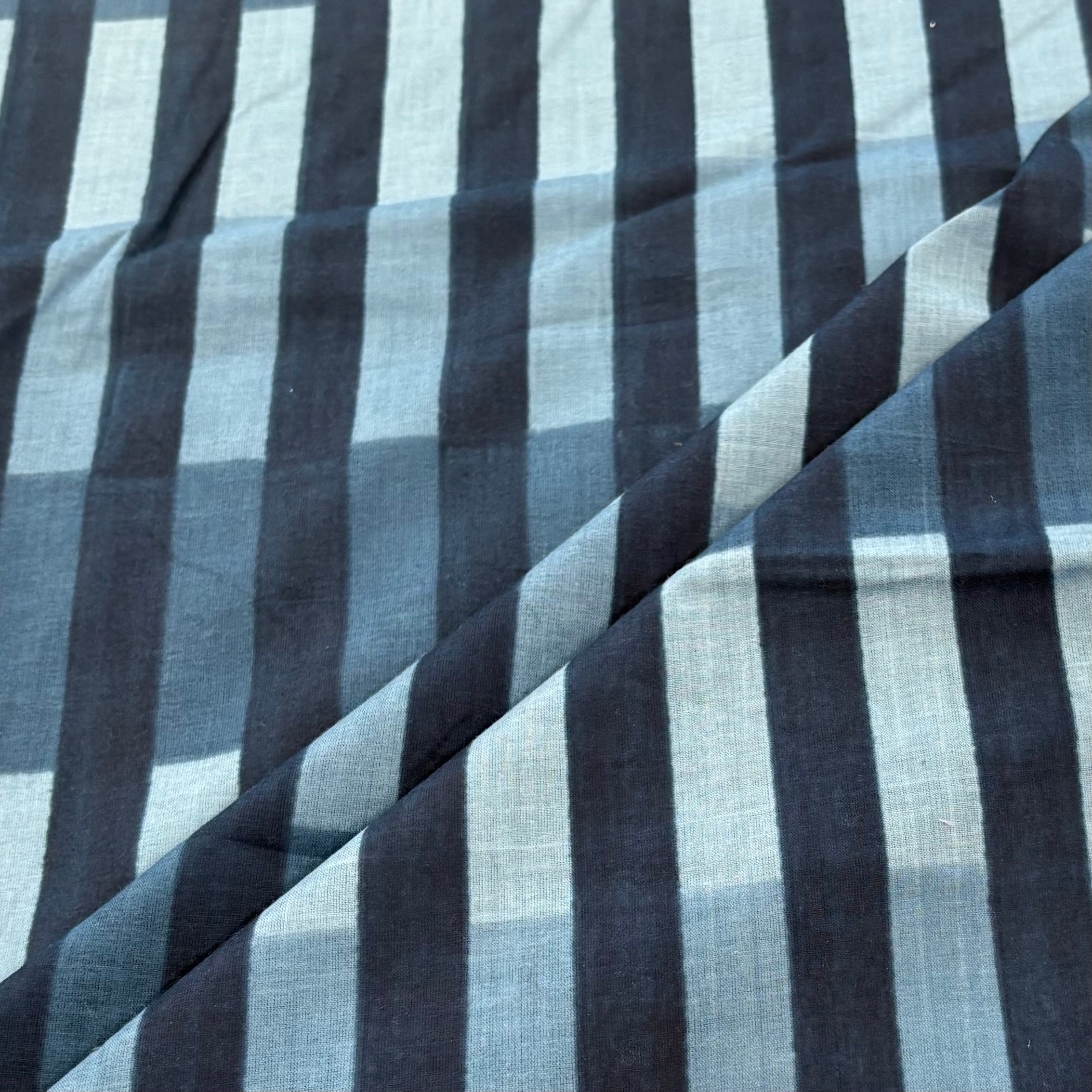 A close-up of a striped fabric with alternating wide and narrow vertical stripes in dark navy, charcoal gray, and light gray, shown with a diagonal fold.