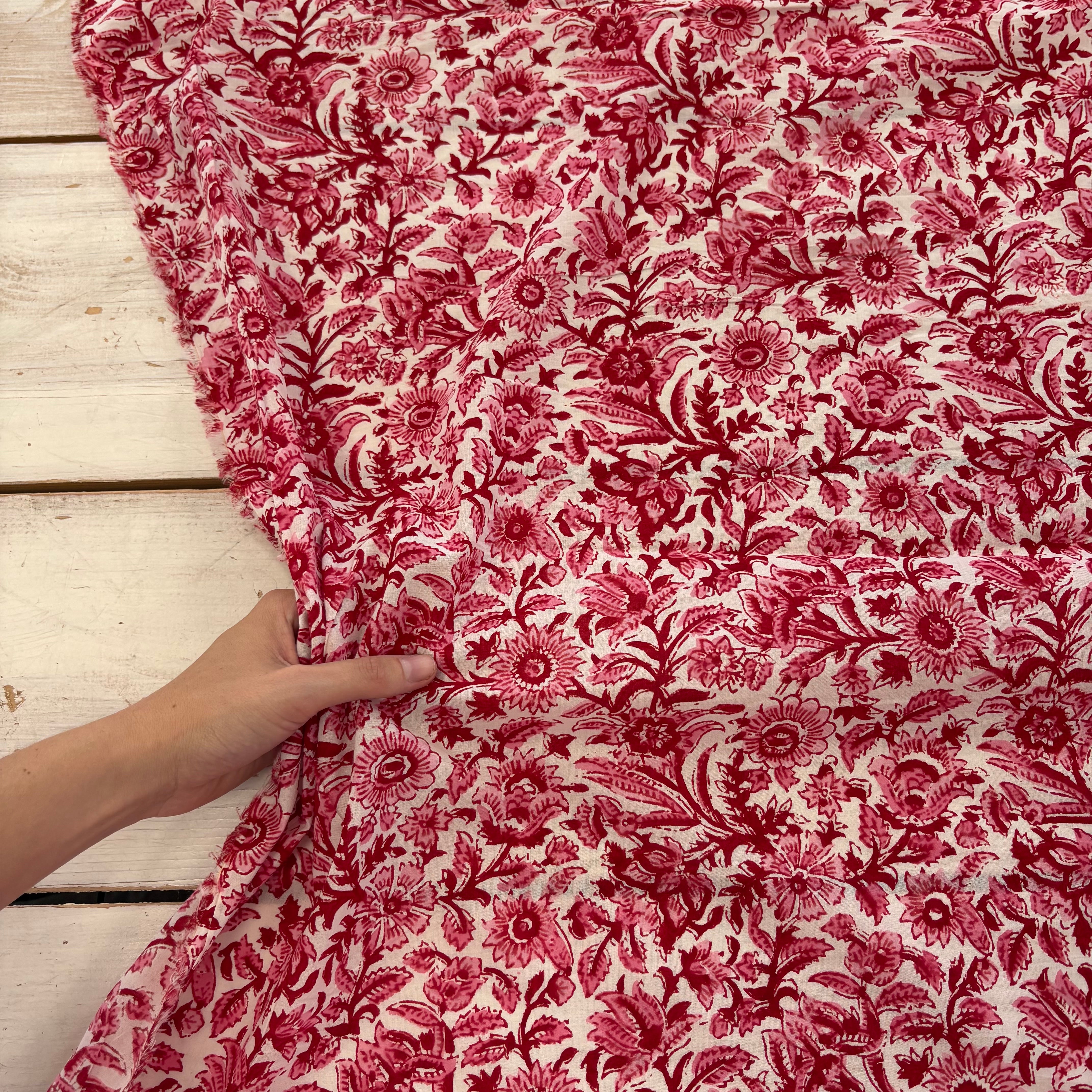 A close-up of a red and pink floral fabric with a hand holding the corner to show its drape and texture, laid over a white wooden plank surface.