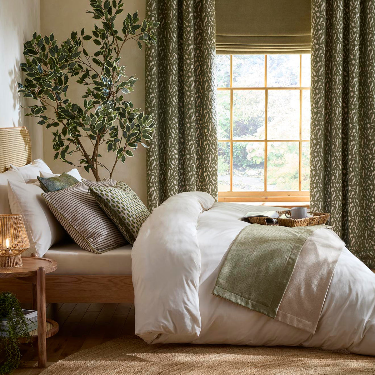 Zafira Cedar bedroom room set. A styled bedroom scene featuring patterned sage green curtains, a wooden bed with white bedding, a side table with a lamp, and a window with a Roman blind.