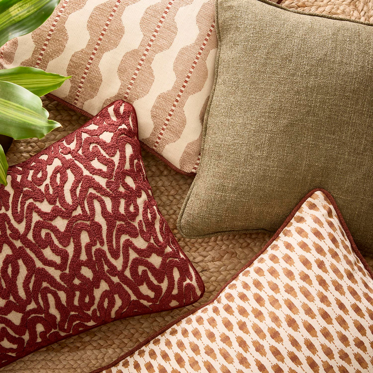 Zafira Cushions Four decorative throw pillows arranged on a woven sofa. One pillow has a deep red abstract pattern on cream, one is solid olive green linen, one has wavy beige stripes on cream, and one has a tan geometric dot pattern on cream.