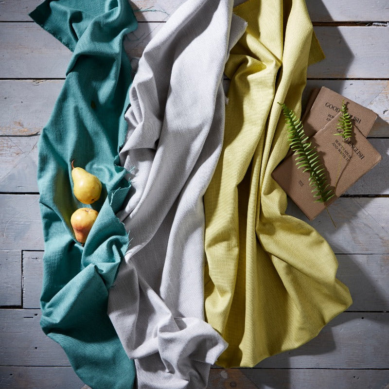 Three folded cloths in teal, white, and yellow on a wooden surface with a small plant.