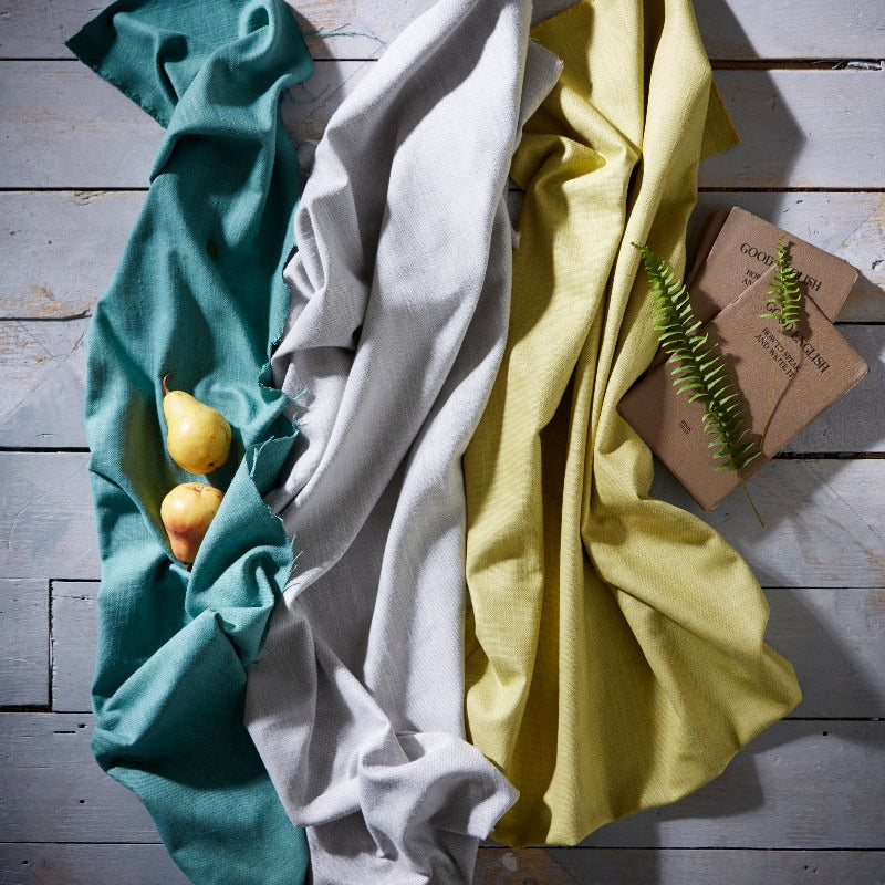 Three folded cloths in teal, white, and yellow on a wooden surface with a small plant.