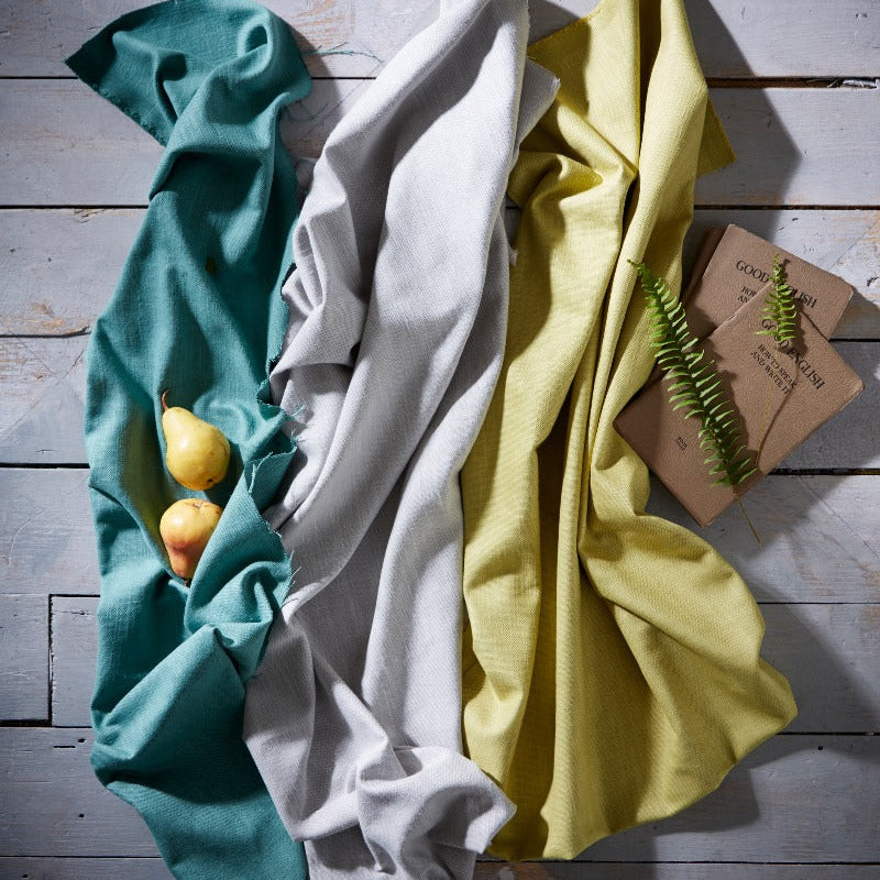 Three folded cloths in teal, white, and yellow on a wooden surface with a small plant.