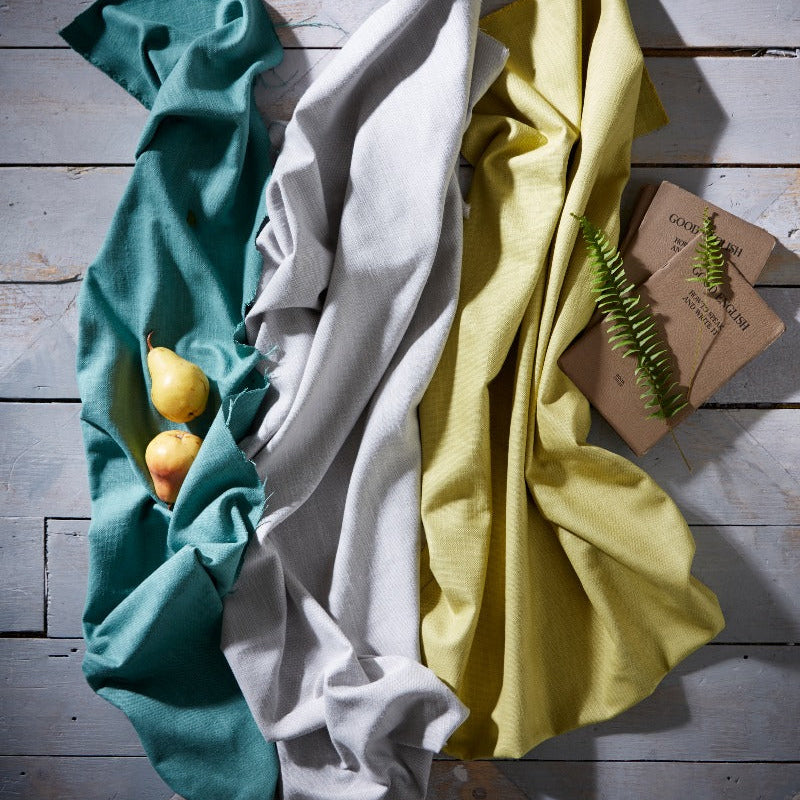 Three folded cloths in teal, white, and yellow on a wooden surface with a small plant.