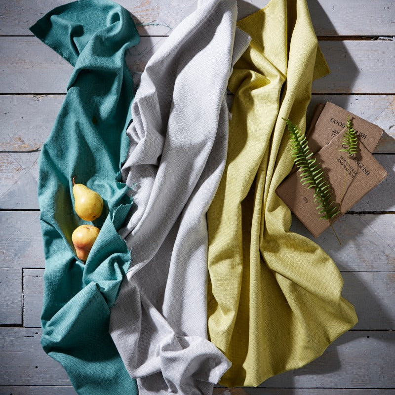 Three folded cloths in teal, white, and yellow on a wooden surface with a small plant.