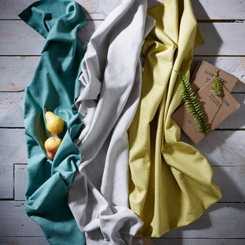 Three folded cloths in teal, white, and yellow on a wooden surface with a small plant.