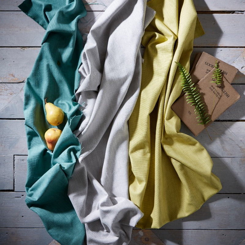 Three folded cloths in teal, white, and yellow on a wooden surface with a small plant.