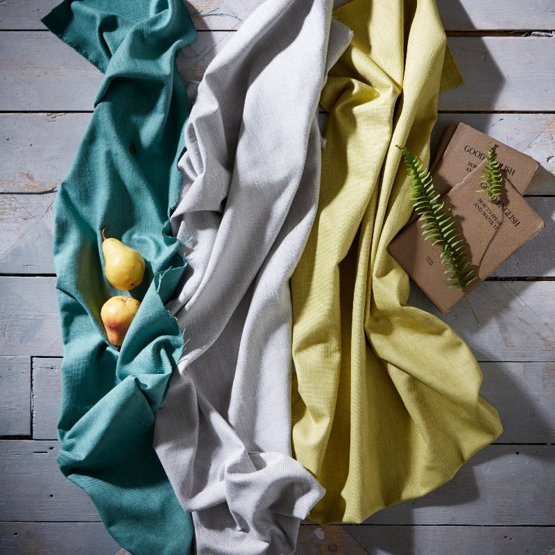 Three folded cloths in teal, white, and yellow on a wooden surface with a small plant.