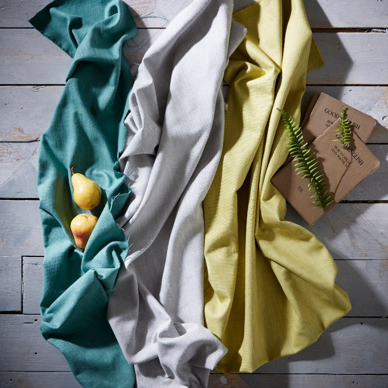 Three folded cloths in teal, white, and yellow on a wooden surface with a small plant.