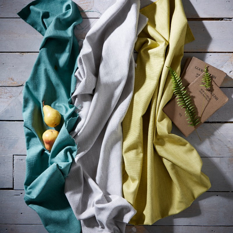 Three folded cloths in teal, white, and yellow on a wooden surface with a small plant.