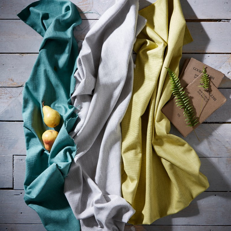 Three folded cloths in teal, white, and yellow on a wooden surface with a small plant.