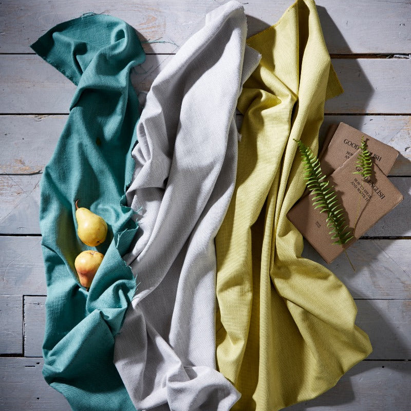 Three folded cloths in teal, white, and yellow on a wooden surface with a small plant.