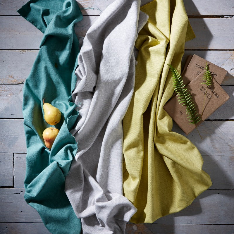 Three folded cloths in teal, white, and yellow on a wooden surface with a small plant.