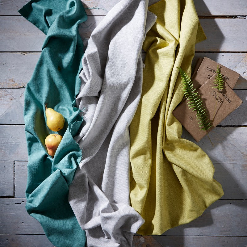 Three folded cloths in teal, white, and yellow on a wooden surface with a small plant.