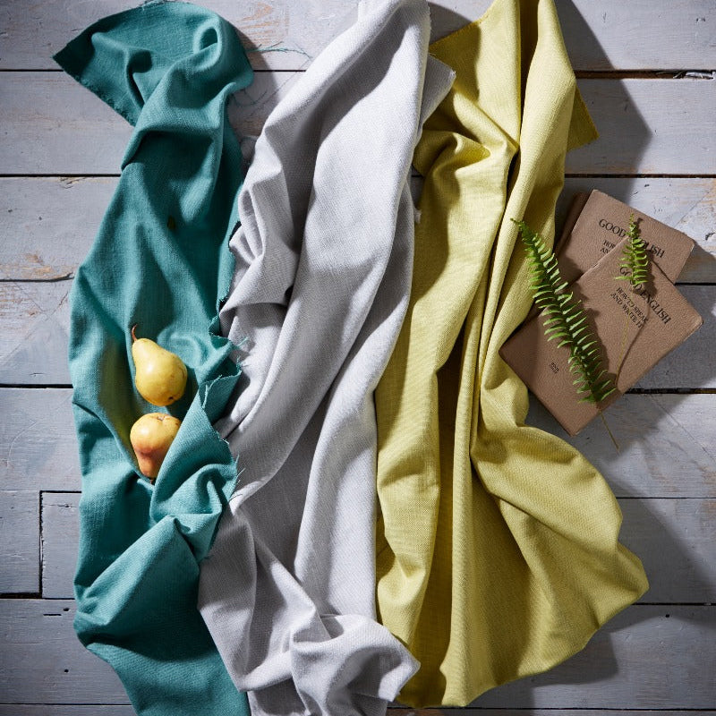 Three folded cloths in teal, white, and yellow on a wooden surface with a small plant.