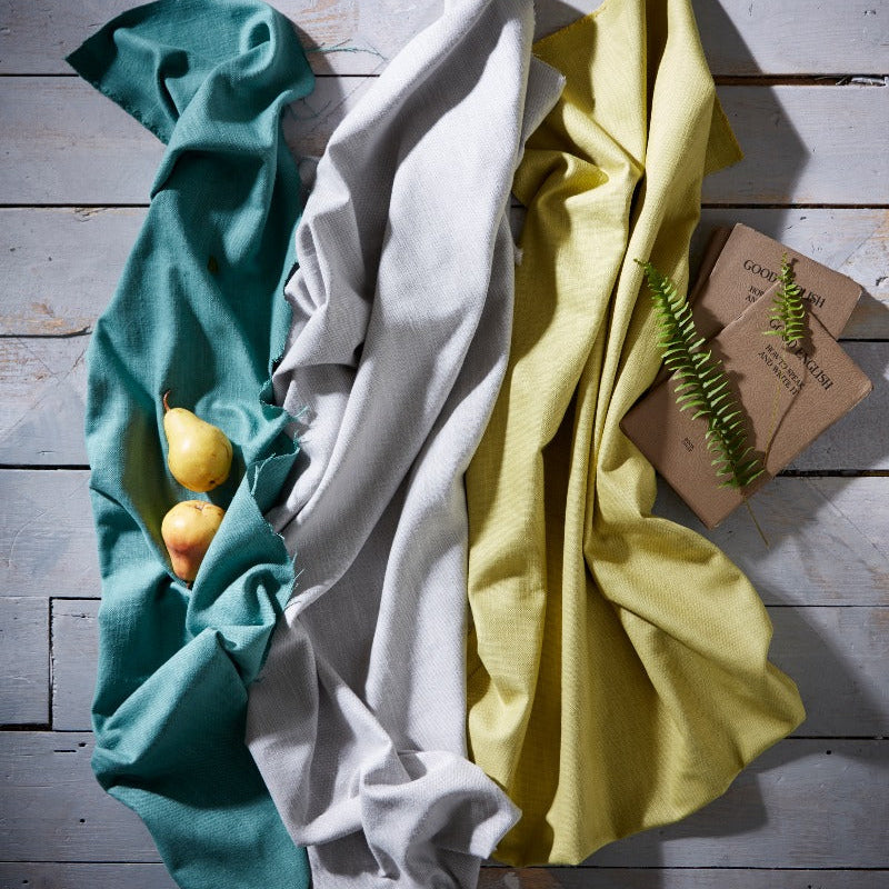 Three folded cloths in teal, white, and yellow on a wooden surface with a small plant.