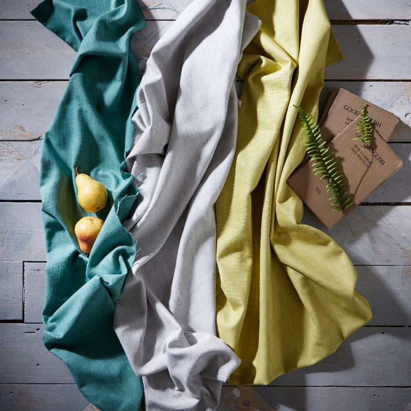 Three folded cloths in teal, white, and yellow on a wooden surface with a small plant.