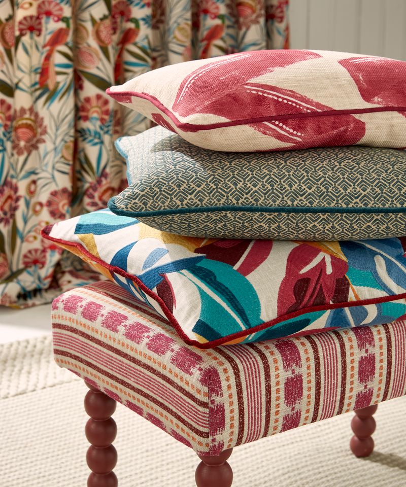 Stack of colorful patterned cushions on a matching ottoman with floral curtains in the background.