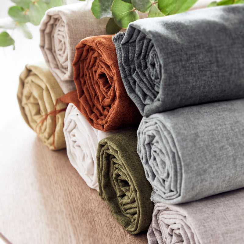 Stack of folded fabric rolls in various colors on a wooden surface with green leaves in the background.