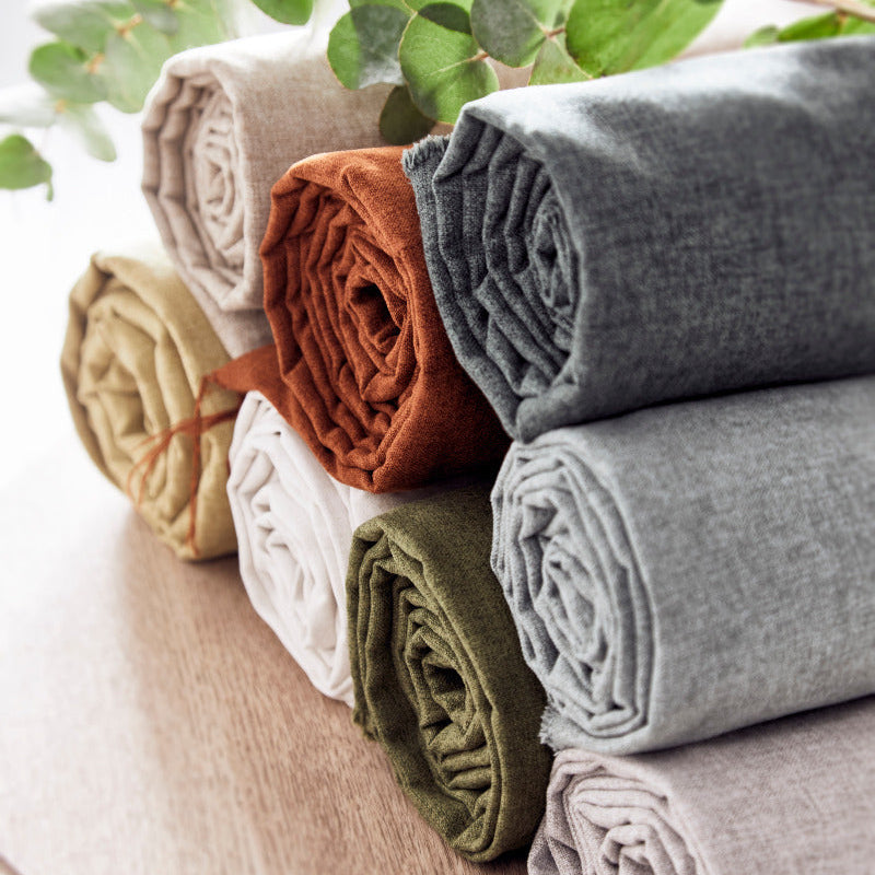Stack of folded fabric rolls in various colors on a wooden surface with green leaves in the background.