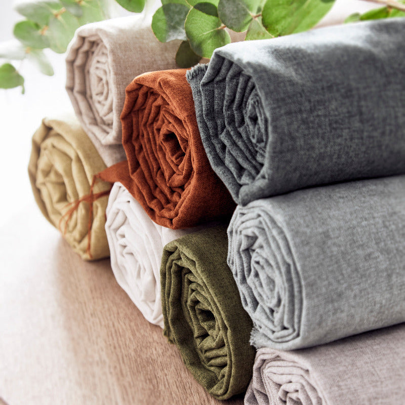 Stack of folded fabric rolls in various colors on a wooden surface with green leaves in the background.