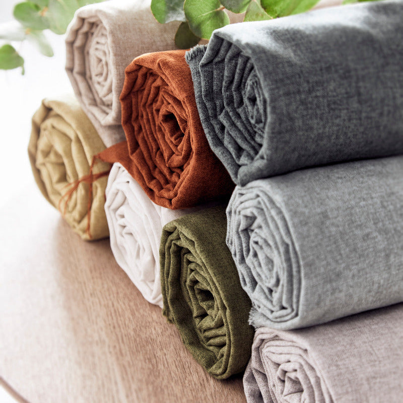 Stack of folded fabric rolls in various colors on a wooden surface with green leaves in the background.