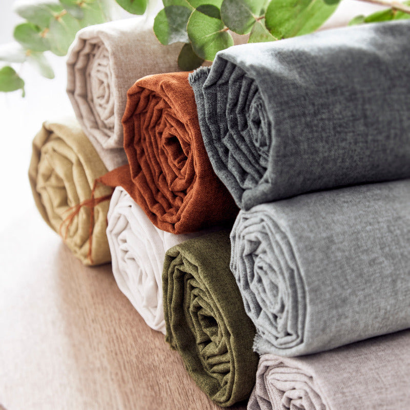 Stack of folded fabric rolls in various colors on a wooden surface with green leaves in the background.