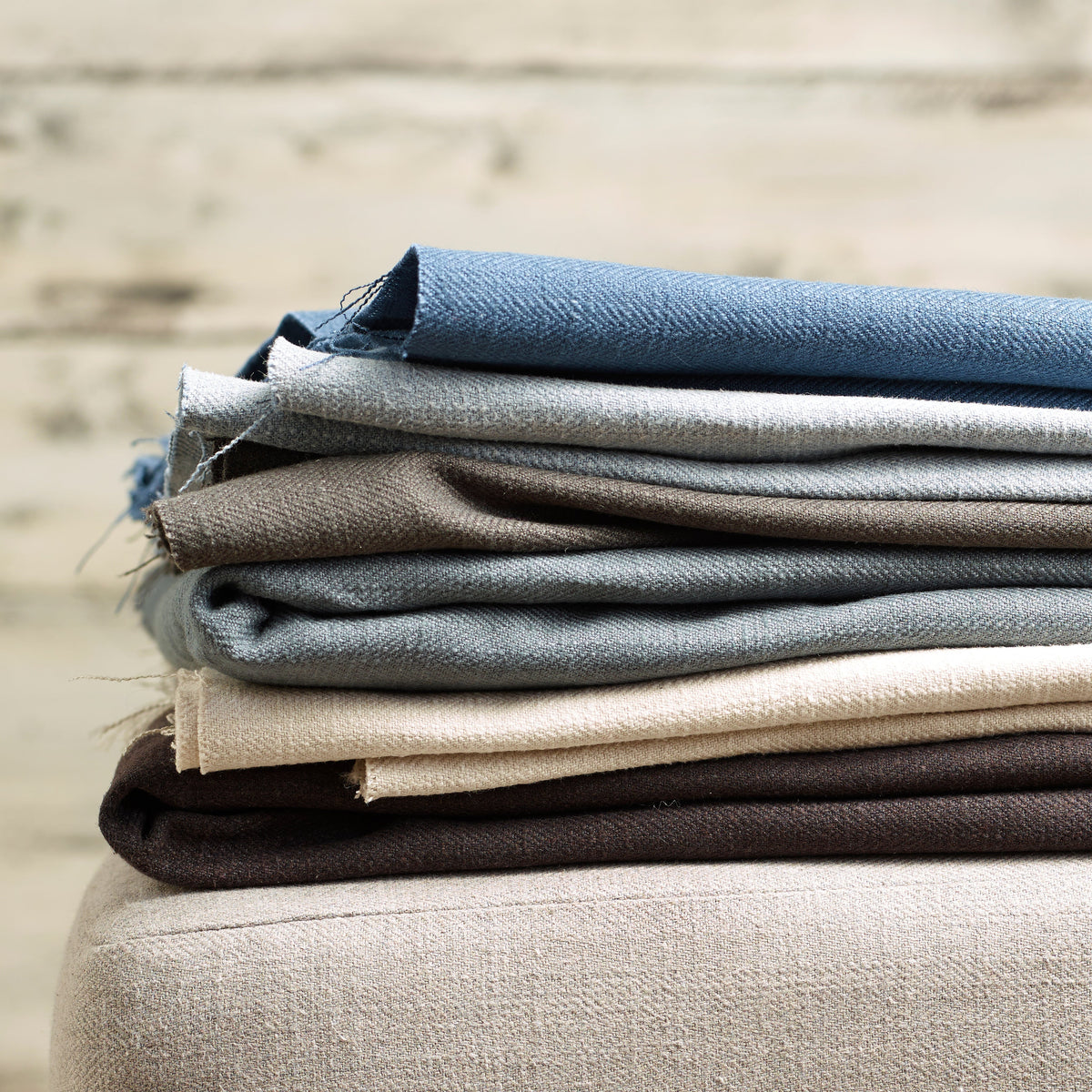 A stack of folded woven fabric swatches in blue, gray, taupe, cream, and dark brown, resting on a light beige upholstered surface against a softly blurred neutral background.