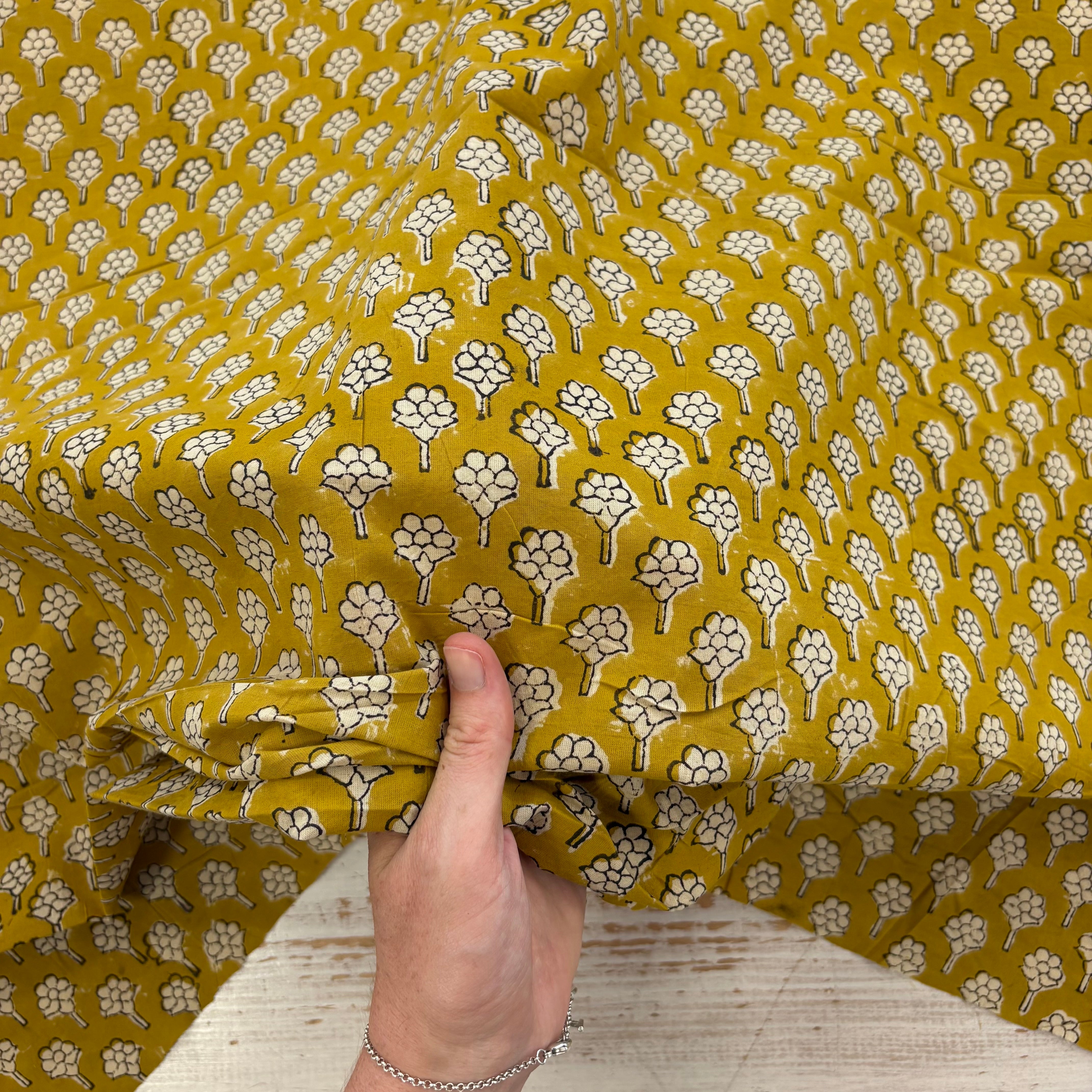 A close-up of a mustard yellow fabric with a repeating white and black floral print, held by a hand to show its texture and drape.