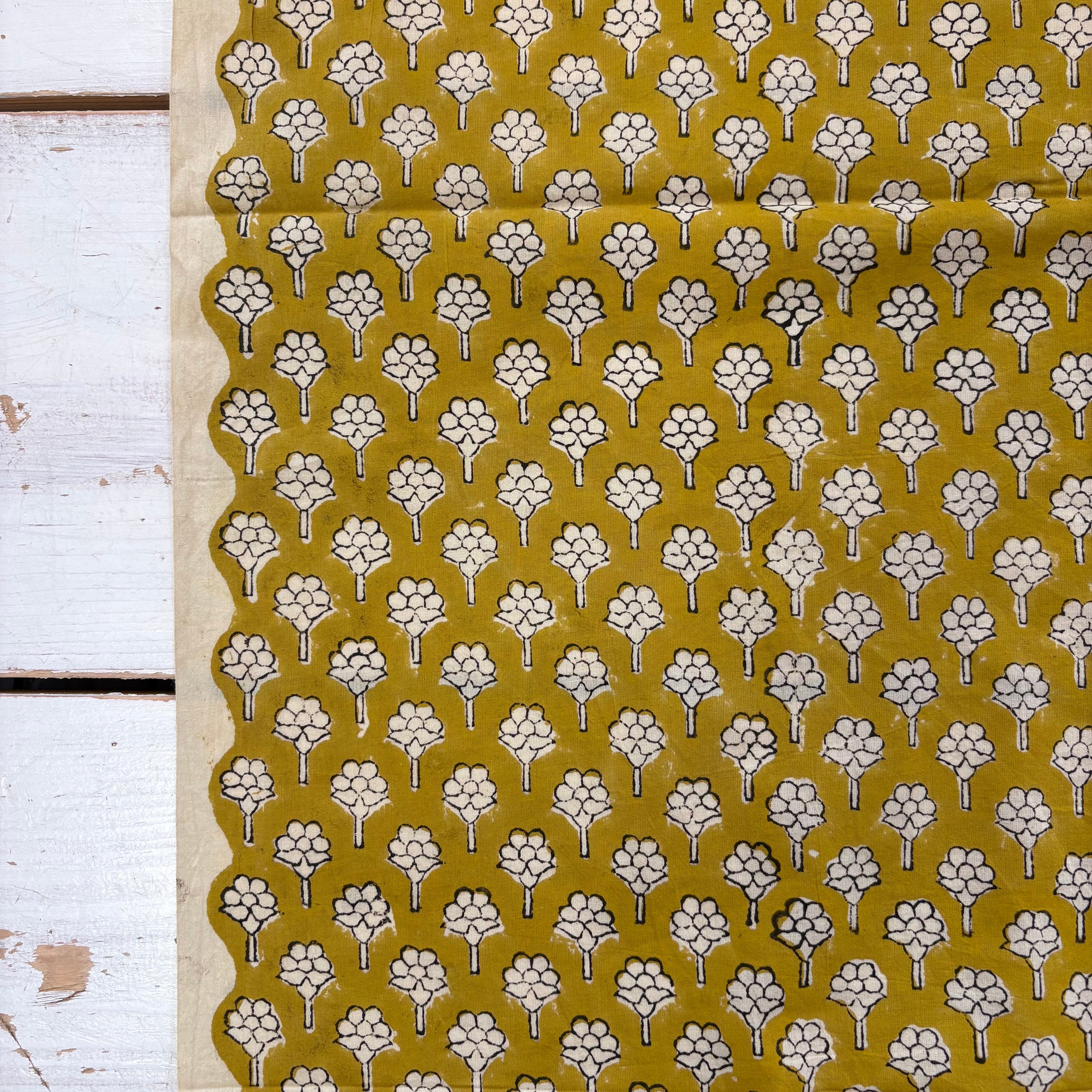 A close-up of a fabric or textile laid flat on a white wooden surface, featuring a repeating floral motif in white and dark brown on a mustard yellow background.
